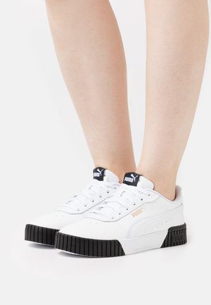 Baskets Puma blanches avec semelles noires portées pieds nus, arborant un logo sur la languette et sur le côté, sur fond blanc.