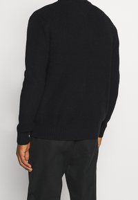 Jersey de punto negro con una textura acanalada, que presenta mangas largas y un escote redondo. La vista trasera muestra un ajuste relajado y sutiles detalles de costura.
