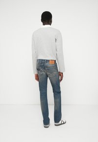 Camisa cinza de mangas longas, combinada com jeans azuis desgastados com bolsos traseiros e um retângulo de couro castanho. Ténis preto e branco completam o visual.