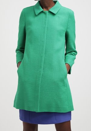 Manteau vert texturé avec un col et des manches longues, doté de poches latérales et d'une coupe droite. Porté sur une jupe bleue.