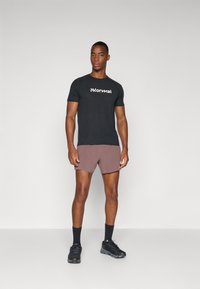 T-shirt noire à manches courtes avec le logo "Normal" en blanc, associée à un short marron clair. Le modèle porte des chaussures de sport noires et des chaussettes sombres.
