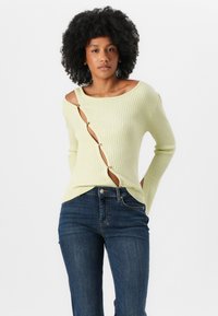 Light yellow ribbed sweater featuring a diagonal button detail and cut-out design, paired with dark blue denim jeans.