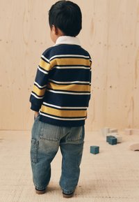 Pull rayé bleu et jaune avec un col blanc, associé à un pantalon cargo en denim bleu ample. L'arrière-plan présente des textures et des blocs en bois.
