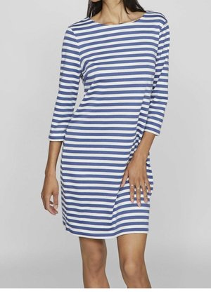 Kvinde iført en blå og hvid kjole med horisontale striber, der går til knæet og har trekvartlange ærmer, stående med den ene hånd hvilede på hoften.