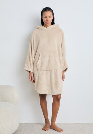 Huvtröja i oversized-modell i mjuk beige tyg med en texturerad, fluffig yta. Har en framficka och avslappnade ärmar som slutar ovanför knäna.