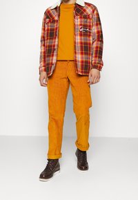 Koszula w kratę flanelową w kolorach czerwonym, brązowym i żółtym z kołnierzem z owczej skóry, pomarańczowa koszulka z okrągłym dekoltem oraz pomarańczowe spodnie sztruksowe. Brązowe buty.