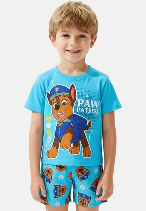 Jeune garçon souriant portant un t-shirt bleu Paw Patrol et un short assorti avec le personnage Chase imprimé sur les deux.