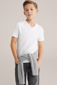 Balti medvilninė V formos kaklo marškinėliai, derinami su pilku megztu mezginiu, surištu aplink juosmenį, ir tamsiai pilkomis džinsinėmis kelnėmis.