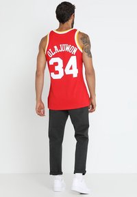 Mann trägt ein rotes Basketball-Trikot mit "Olajuwon 34" auf dem Rücken, dunkle Hose, weiße Turnschuhe, steht vor einem weißen Hintergrund, mit dem Rücken zur Kamera.