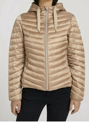Kobieta ubrana w jasnobrązową pikowaną kurtkę z kapturem, zapinaną na zamek z przodu i ze sznurkami, w połączeniu z czarnymi spodniami.