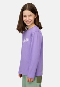 Langärmliges lila Baumwoll-T-Shirt mit einem weißen Tiergraphic auf der Vorderseite, kombiniert mit grünen Hosen, präsentiert von einem lächelnden Kind.