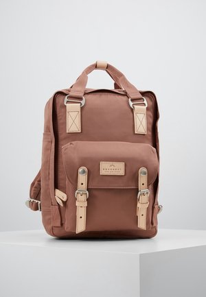 Sac à dos carré rose poudré avec des sangles en cuir, une poche avant avec fermetures à boucle, et des poignées supérieures sur une surface blanche.