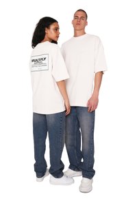 Camiseta de corte relajado en color crema con gráfico negro en la parte de atrás, combinada con jeans azules. Modelo lleva zapatillas blancas, mostrando un look casual.