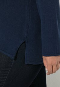 Laivastonsininen neulottu neulepaita, jossa on teksturoitu pinta, pitkät hihat ja sivuhalkiot helmassa, puettuna tummien farkkujen kanssa.