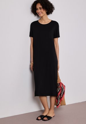 Kvinde iført en sort midi-kjole med korte ærmer og sorte slidesandaler, der holder en brun taske med et rødt og leopardprintet tørklæde.