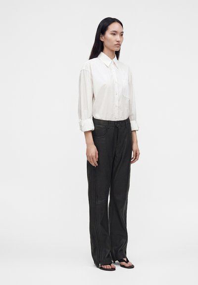 Femme aux longs cheveux foncés porte une chemise blanche boutonnée avec les manches retroussées, un pantalon noir à jambes larges avec des détails zippés, et des sandales noires à bouts ouverts.