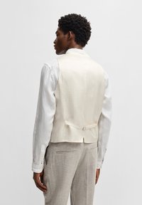 Cremfarbener Polyester-Westen mit strukturiertem Oberflächen, taillierter Schnitt, verstellbare Rückenstrippe, getragen über einem hellen Hemd und grauen Hosen.