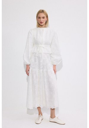 Γυναίκα με ξανθά μαλλιά που φοράει λευκό μακρυμάνικο φόρεμα με φουσκωτά μανίκια και λευκά δερμάτινα παπούτσια με κορδόνια, που στέκεται μπροστά σε λευκό φόντο.