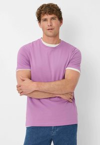 T-shirt viola chiaro con collo rotondo e bordo bianco, maniche corte, realizzata in tessuto morbido. Indossata con jeans in denim blu.