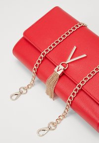 Červené texturované psaníčko se zlatým řetízkovým popruhem, zdobené střapcem a zlatým zapínáním s akcentem ve tvaru písmena 'V'.