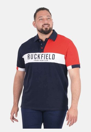 Homme portant un polo bleu marine et rouge avec le texte "Ruckfield French Rugby Club", debout et regardant légèrement vers le haut et la droite.