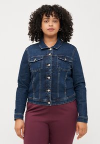 Ženska s kodrastimi lasmi nosi prilegajočo se jeans jakno in bordo hlače. Stoji ob običajni beli ozadju in gleda neposredno v kamero.