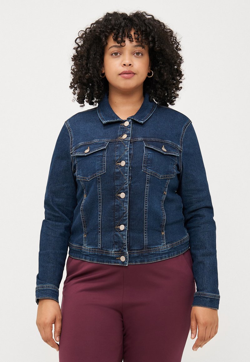 Ženska s kodrastimi lasmi nosi prilegajočo se jeans jakno in bordo hlače. Stoji ob običajni beli ozadju in gleda neposredno v kamero.