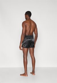 Calções de cetim preto com cintura franzida, aberturas laterais e textura suave, mostrados de costas num modelo descalço.