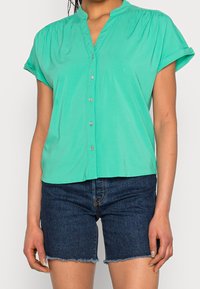 Blusa verde de manga corta con cuello, cierre de botones en la parte delantera y detalles fruncidos en los hombros, combinada con pantalones cortos de mezclilla oscura.