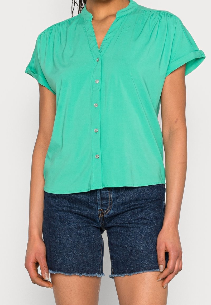 Blusa verde de manga corta con cuello, cierre de botones en la parte delantera y detalles fruncidos en los hombros, combinada con pantalones cortos de mezclilla oscura.