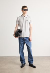 Polo cinza claro com detalhes em preto e logotipo 'BOSS', jeans de denim azul, sapatos pretos e óculos de sol oversized pretos, segurando uma bolsa preta.