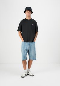 Zwarte katoenen t-shirt met logo, lichtblauwe denim shorts en zwarte bucket hat. Witte en zwarte sneakers maken de outfit compleet.