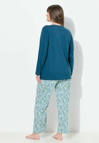 Kvinde stående barfodet med ansigtet mod en hvid væg, iført en teal langærmet bluse og hvide pyjamasbukser med blå og gule blomstrede striber.