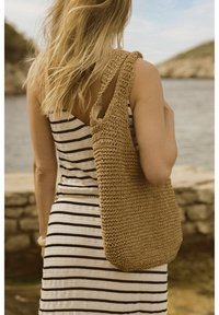 Borsa tote in paglia naturale intrecciata con una singola tracolla, superficie texture e colore marrone sabbioso; indossata sopra un vestito a righe bianco e nero.