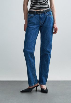 Kvinde iført højtaljede, lige blå jeans, sort bælte, stribet ærmeløs top og sorte spidse hælede sko, stående på gråt gulv.