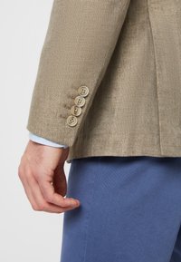 Linen blejzer v hnedej farbe s štyrmi krémovými gombíkmi na rukáve. Vnútro obsahuje modrú košeľu viditeľnú pod manžetou.