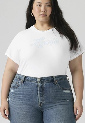 Kvinde med langt mørkt hår iført hvid Levi’s t-shirt og højttaljede blå jeans, stående foran en ensfarvet grå baggrund.