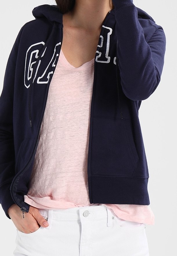 Sweat à capuche zippé bleu marine avec l'inscription "GAP" en blanc, fait en tissu doux. Porté par-dessus un T-shirt col en V rose clair, associé à un pantalon blanc.