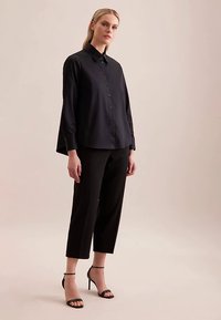 Camicia nera con bottoni, maniche lunghe e vestibilità comoda, abbinata a pantaloni neri corti e tacchi alti con cinturini. Tessuto dalla texture liscia.