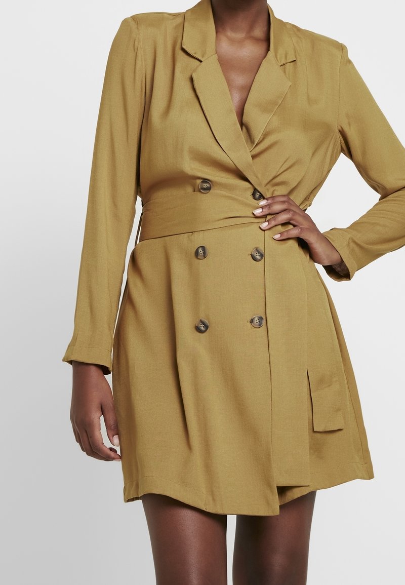 Femme portant une robe blazer jaune moutarde double boutonnage avec manches longues et une poche latérale, main sur la hanche, sur un fond uni.