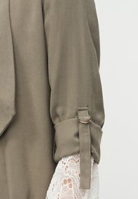 Manche en tissu couleur taupe avec un revers roulé maintenu par une sangle et une boucle, sur un vêtement en dentelle blanche en dessous.