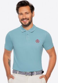 Jasnoniebieska koszulka polo o ryflowanej teksturze, z trzema guzikami przy dekolcie i czerwonym emblematem na piersi. W zestawieniu z wzorzastym paskiem.