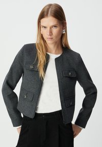 Szara kurtka z wełny z okrągłym dekoltem, przednim zamkiem, dwoma kieszeniami na piersi oraz ozdobnymi metalowymi akcentami. Noszona na białej koszuli.