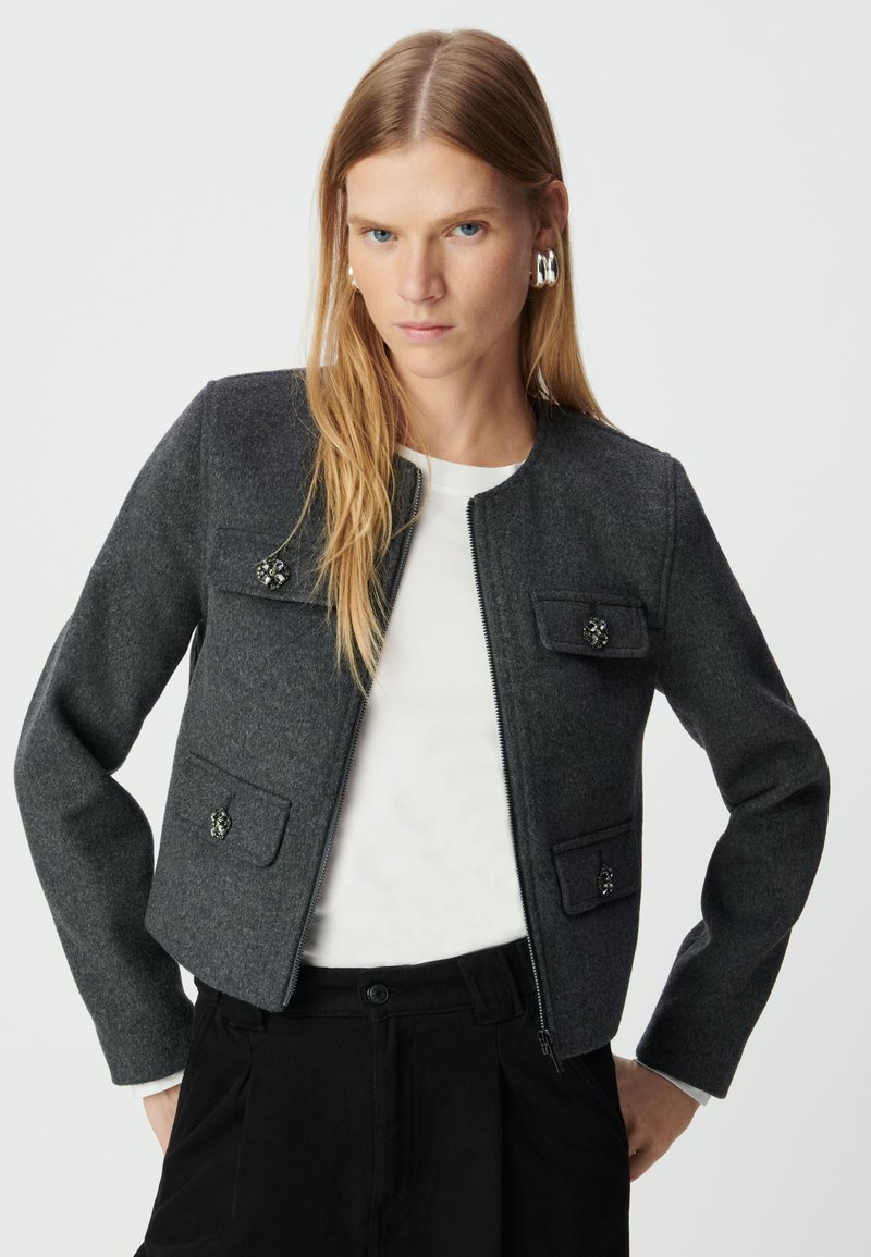 Szara kurtka z wełny z okrągłym dekoltem, przednim zamkiem, dwoma kieszeniami na piersi oraz ozdobnymi metalowymi akcentami. Noszona na białej koszuli.