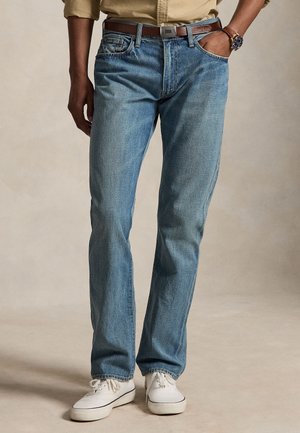 Mand iført lyseblå jeans med lige ben, hvide sneakers, brun læderbælte, beige skjorte og armbåndsur, stående med hænderne ned langs siden.