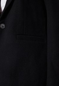 Blazer nero con una texture liscia, caratterizzato da una chiusura con un solo bottone e una tasca laterale. Il tessuto appare morbido e leggermente opaco.