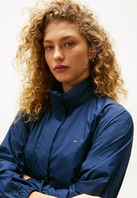 Granatowa kurtka wiatrówka z wysokim kołnierzem, elastycznymi mankietami i małym akcentem w postaci logo. Wykonana z gładkiego, lekkiego materiału.