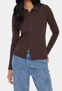 Brown, ribbed long-sleeve button-up shirt with a collar, features a fitted design and side slits, paired with blue high-waisted jeans.