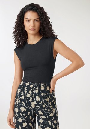 Kobieta o kręconych włosach, ubrana w czarny top bez rękawów i wysokie spodnie z czarnym beżowym kwiatowym wzorem, stoi z jedną ręką na biodrze.