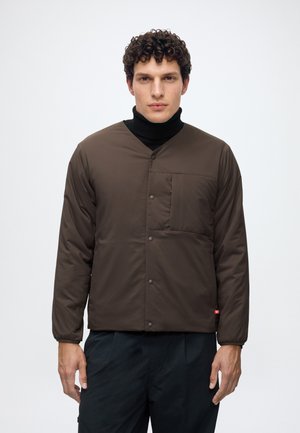 Homme portant une veste matelassée marron foncé avec boutons-pression, une poche poitrine, par-dessus un col roulé noir, debout devant un fond clair uni.